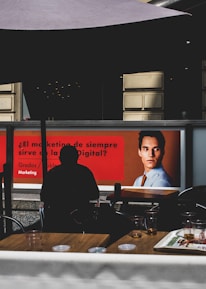 The image depicts a modern indoor setting with posters on a wall featuring a professional headshot and text related to marketing. Silhouetted figures are seated at tables in the foreground, casting shadows. The lighting is dim, creating contrast between the illuminated posters and darker seating area.