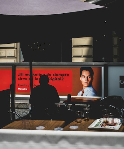 The image depicts a modern indoor setting with posters on a wall featuring a professional headshot and text related to marketing. Silhouetted figures are seated at tables in the foreground, casting shadows. The lighting is dim, creating contrast between the illuminated posters and darker seating area.