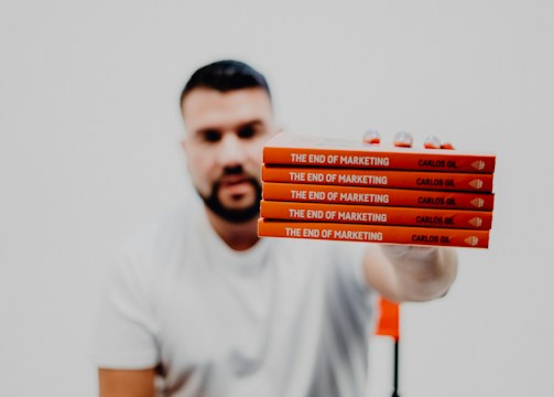 A person in a white t-shirt is holding a stack of five identical red books titled 'The End of Marketing' by Carlos Gil. The background is a very light color, providing minimal distraction from the foreground.