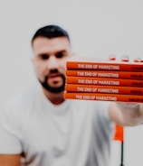 A person in a white t-shirt is holding a stack of five identical red books titled 'The End of Marketing' by Carlos Gil. The background is a very light color, providing minimal distraction from the foreground.