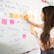 A woman with long hair is writing on a whiteboard. She is using sticky notes and markers to organize and plan a strategy. The board has various handwritten notes and symbols related to marketing and digital education.