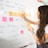 A woman with long hair is writing on a whiteboard. She is using sticky notes and markers to organize and plan a strategy. The board has various handwritten notes and symbols related to marketing and digital education.