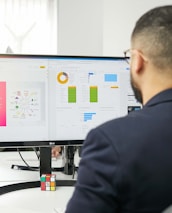 A man in a suit is working at a desk with a dual-monitor setup showing SEO presentation slides and data charts. The office environment is bright, with natural light coming from a nearby window, and a Rubik's cube is placed in front of the monitor.