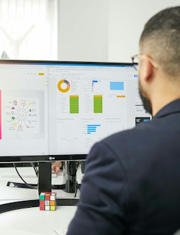 A man in a suit is working at a desk with a dual-monitor setup showing SEO presentation slides and data charts. The office environment is bright, with natural light coming from a nearby window, and a Rubik's cube is placed in front of the monitor.