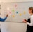Two people are standing in front of a whiteboard that has a weekly schedule for social media content. The board is divided into sections for Twitter, TikTok, Instagram, and Facebook, with sticky notes indicating different content themes for each day of the week. One person is pointing at the board, while the other holds a red marker.