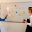 Two people are standing in front of a whiteboard that has a weekly schedule for social media content. The board is divided into sections for Twitter, TikTok, Instagram, and Facebook, with sticky notes indicating different content themes for each day of the week. One person is pointing at the board, while the other holds a red marker.