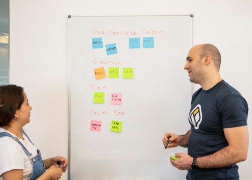 Two people stand in front of a whiteboard covered with colorful sticky notes. The notes are arranged into categories: 'User-Generated Content,' 'Engagement,' 'Event,' and 'Social Wall.' The individuals appear to be in discussion, with one holding a marker.