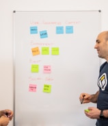 Two people stand in front of a whiteboard covered with colorful sticky notes. The notes are arranged into categories: 'User-Generated Content,' 'Engagement,' 'Event,' and 'Social Wall.' The individuals appear to be in discussion, with one holding a marker.
